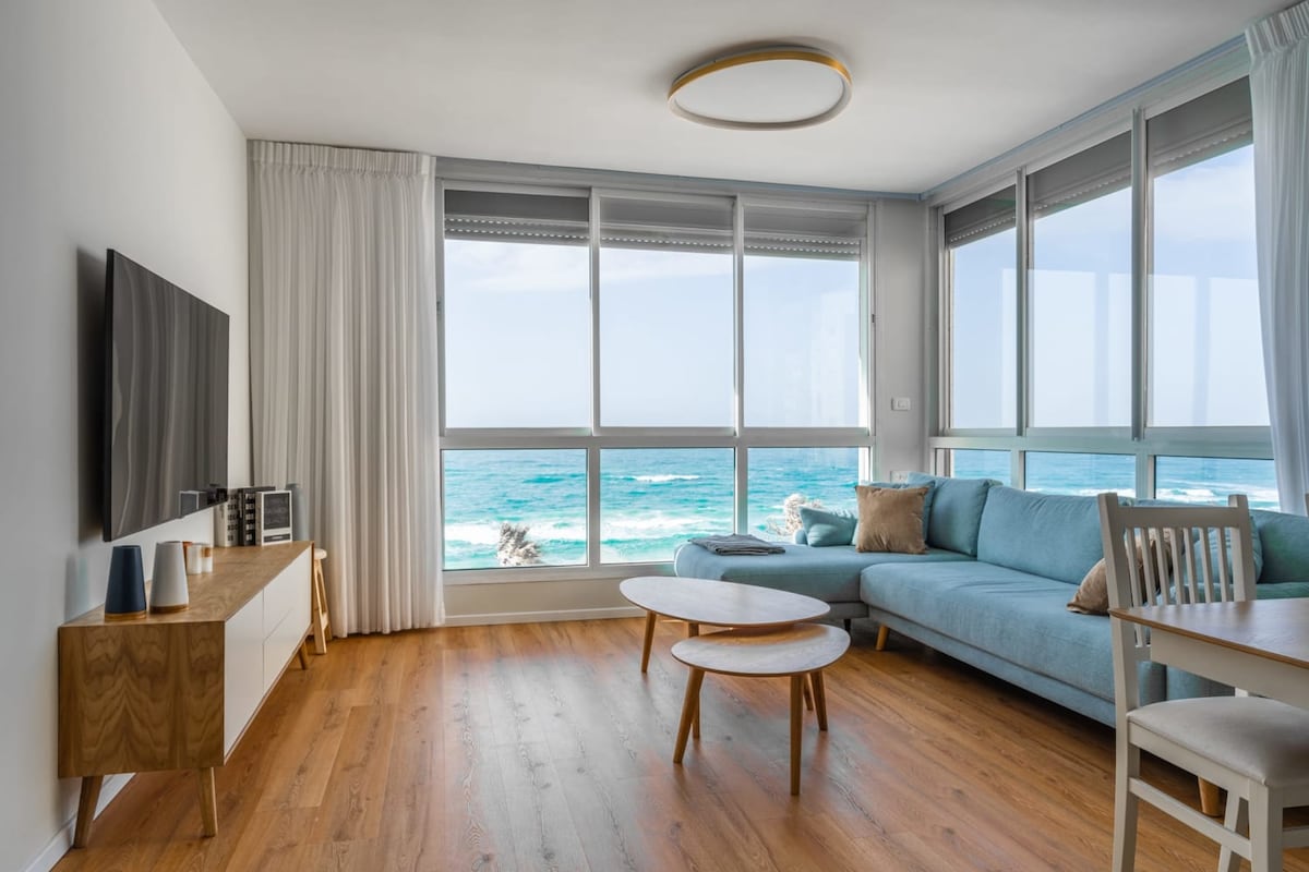 A bright living room showcases panoramic windows with views of the Mediterranean Sea. Light wood flooring extends throughout, complemented by a blue sectional sofa and a low coffee table. A television is mounted on the wall, and sheer curtains add softness to the space.