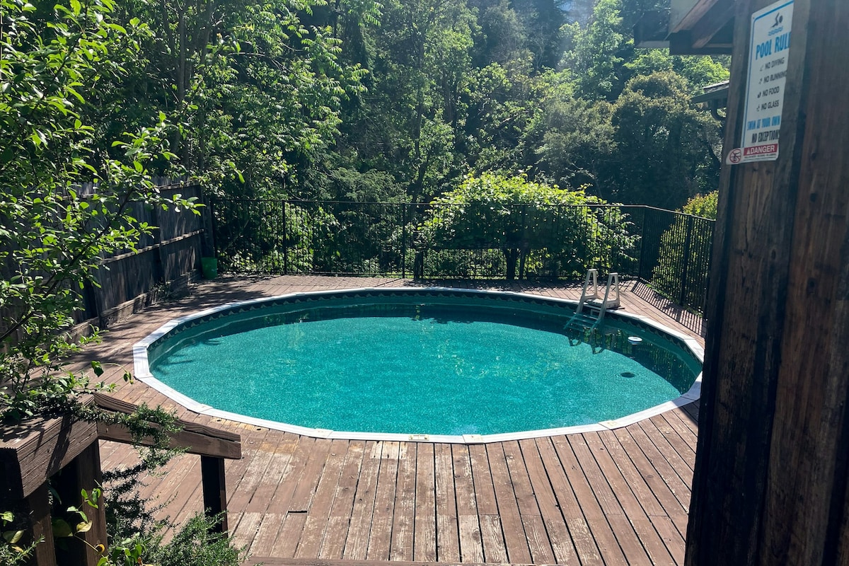 A circular pool is framed by wooden decking, surrounded by lush greenery. Sunlight reflects off the clear water, creating a serene atmosphere. A ladder offers access into the pool, while the natural landscape enhances the sense of tranquility.