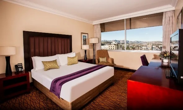 The well-appointed suite features a spacious king bed with decorative pillows. A plush armchair is positioned by the window, providing a view of the surrounding area. A flat-screen TV and a wooden desk with a chair are located nearby, offering convenience for both relaxation and work.