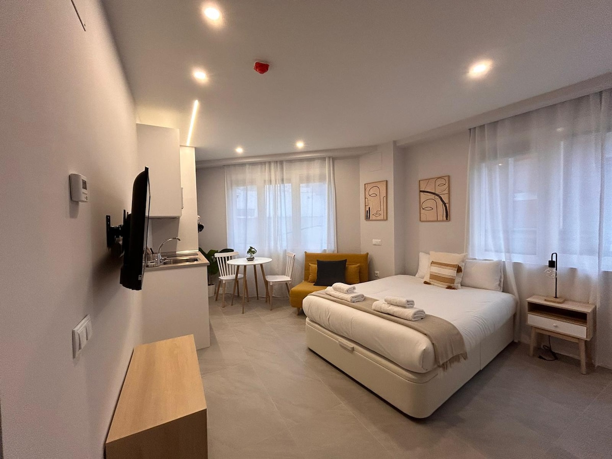 A modern studio space is equipped with a comfortable double bed, a sleek integrated kitchen, and a cozy dining area. Natural light filters through sheer curtains, enhancing the clean design. A small seating area includes an armchair, creating an inviting environment.