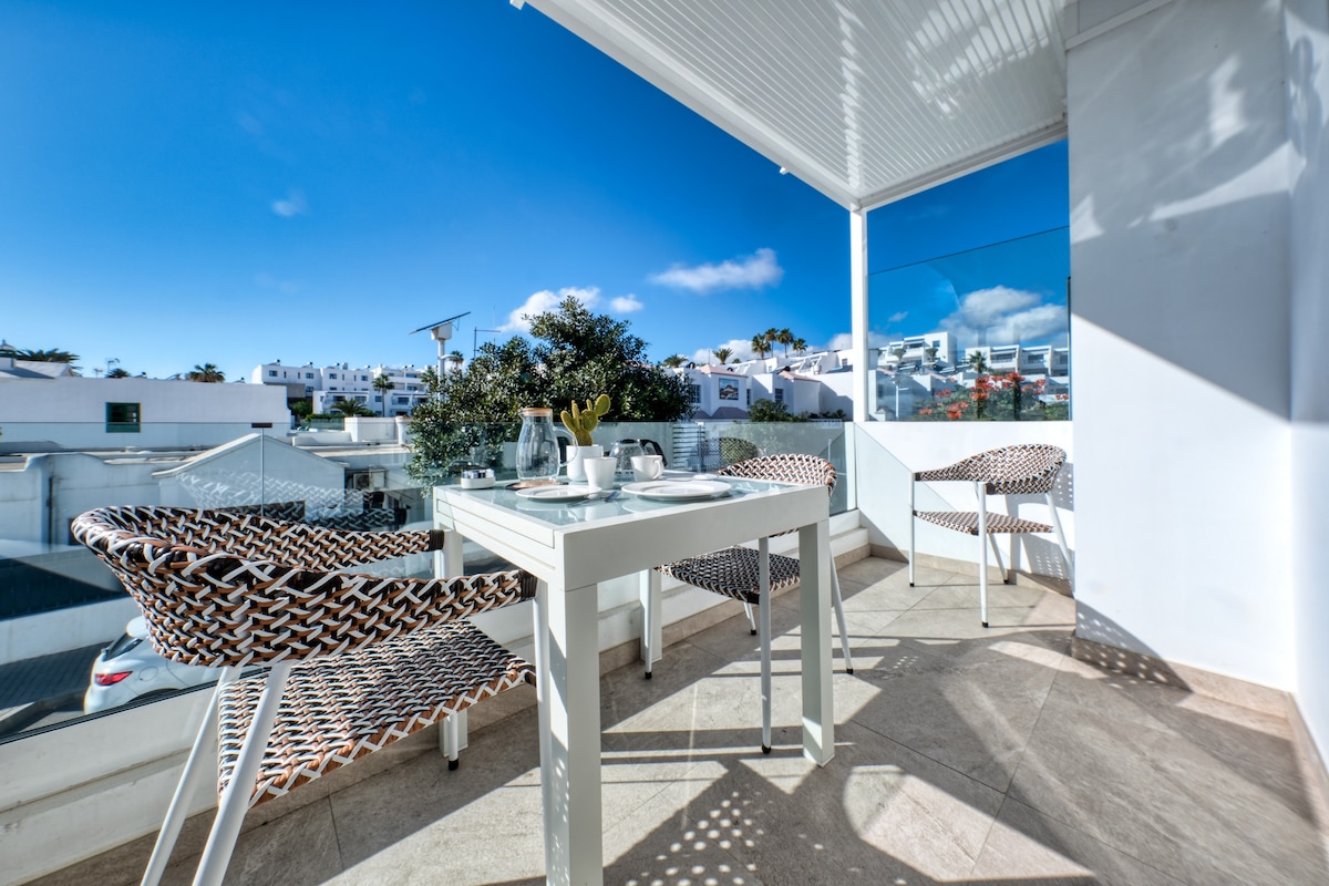 A sunlit terrace features a white table surrounded by four woven chairs. The floor is tiled, and the view includes distant rooftops and clear blue skies. Sunlight creates natural shadows, enhancing the inviting outdoor space.