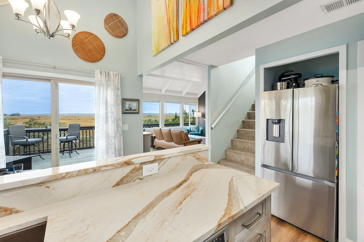 A modern kitchen area features marble countertops and stainless steel appliances, including a refrigerator. Light floods the space from the adjacent living area and outdoor deck, framing views of the expansive marsh. Stairs lead to an upper level, enhancing the home's open layout.