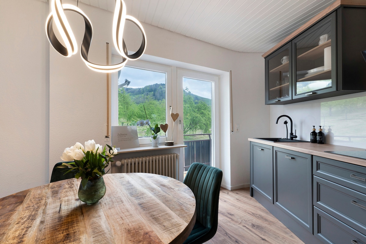 A dining area is presented with a round wooden table, surrounded by upholstered chairs. A modern light fixture hangs above, illuminating the space. Large windows offer a view of the greenery outside, enhancing natural light within the kitchen.