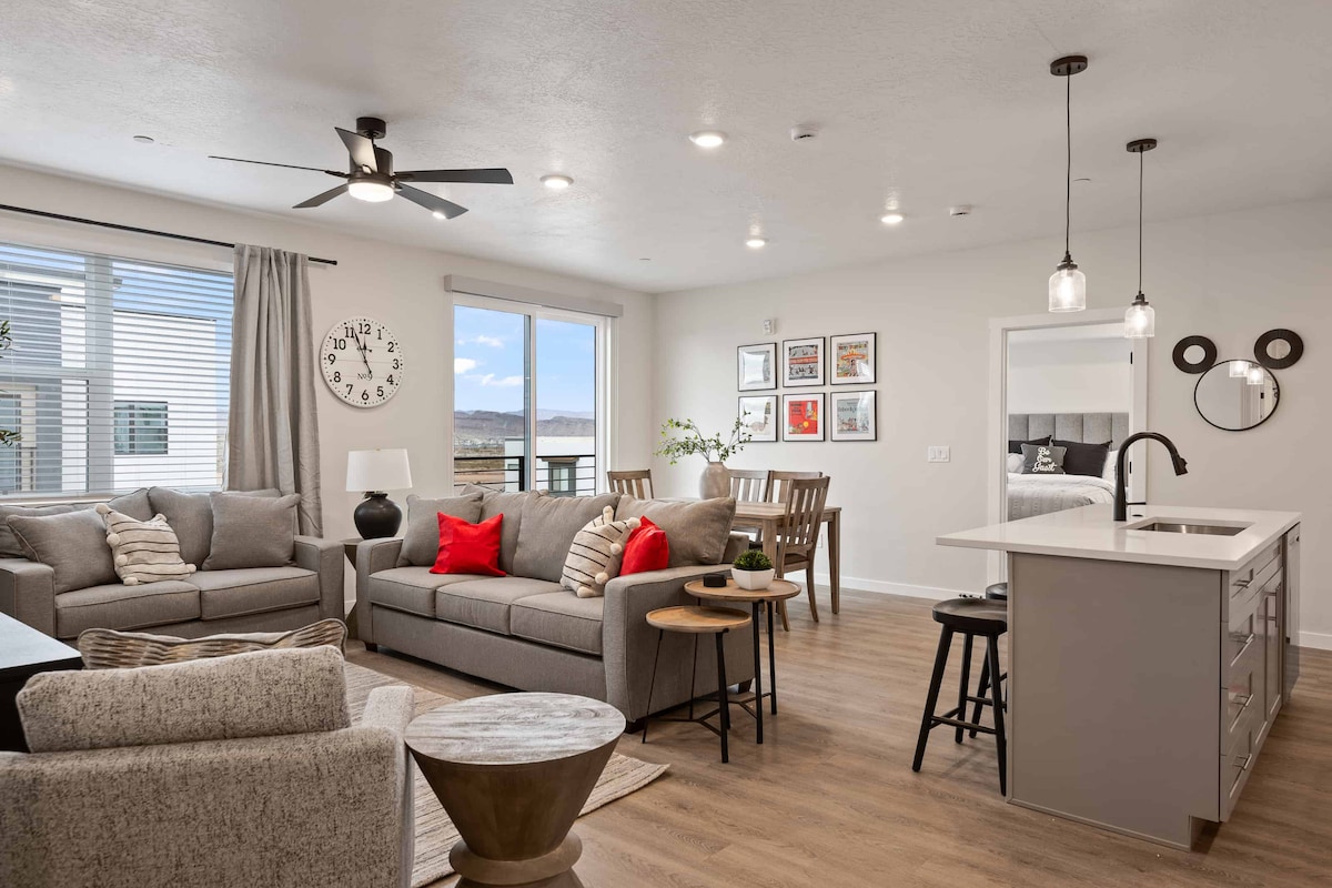 An inviting living area features a light color palette with soft grey sofas adorned with red cushions. A dining table for six is situated near large windows that allow natural light. Stylish artwork decorates the walls, and a modern kitchen island is equipped for convenience.