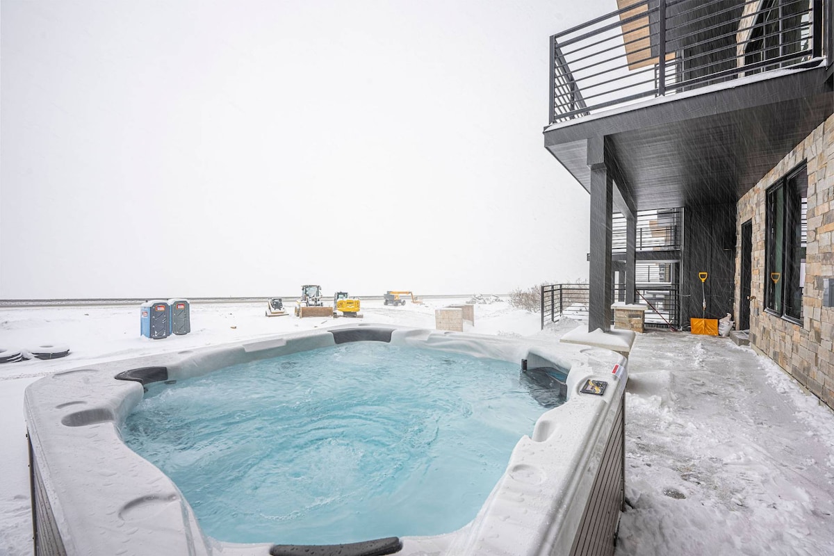 A private outdoor hot tub is visible on a snowy patio, surrounded by a sleek structure with large windows. The scene highlights a winter landscape blanketed in fresh snow, creating a serene atmosphere for relaxation.