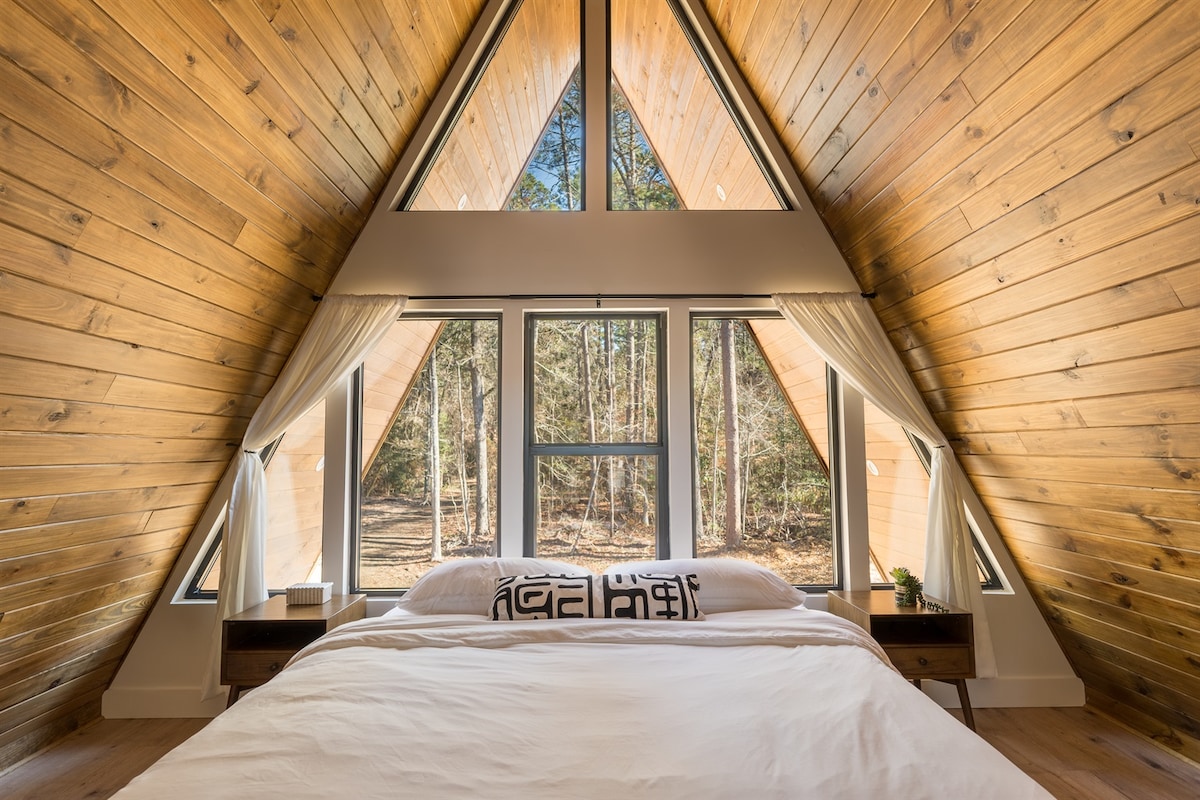 A spacious bedroom is illuminated by natural light streaming through large triangular windows. The room features light wood paneling and a comfortable bed adorned with neutral bedding. Small nightstands flank the bed, contributing to a serene ambiance with views of the surrounding woods.