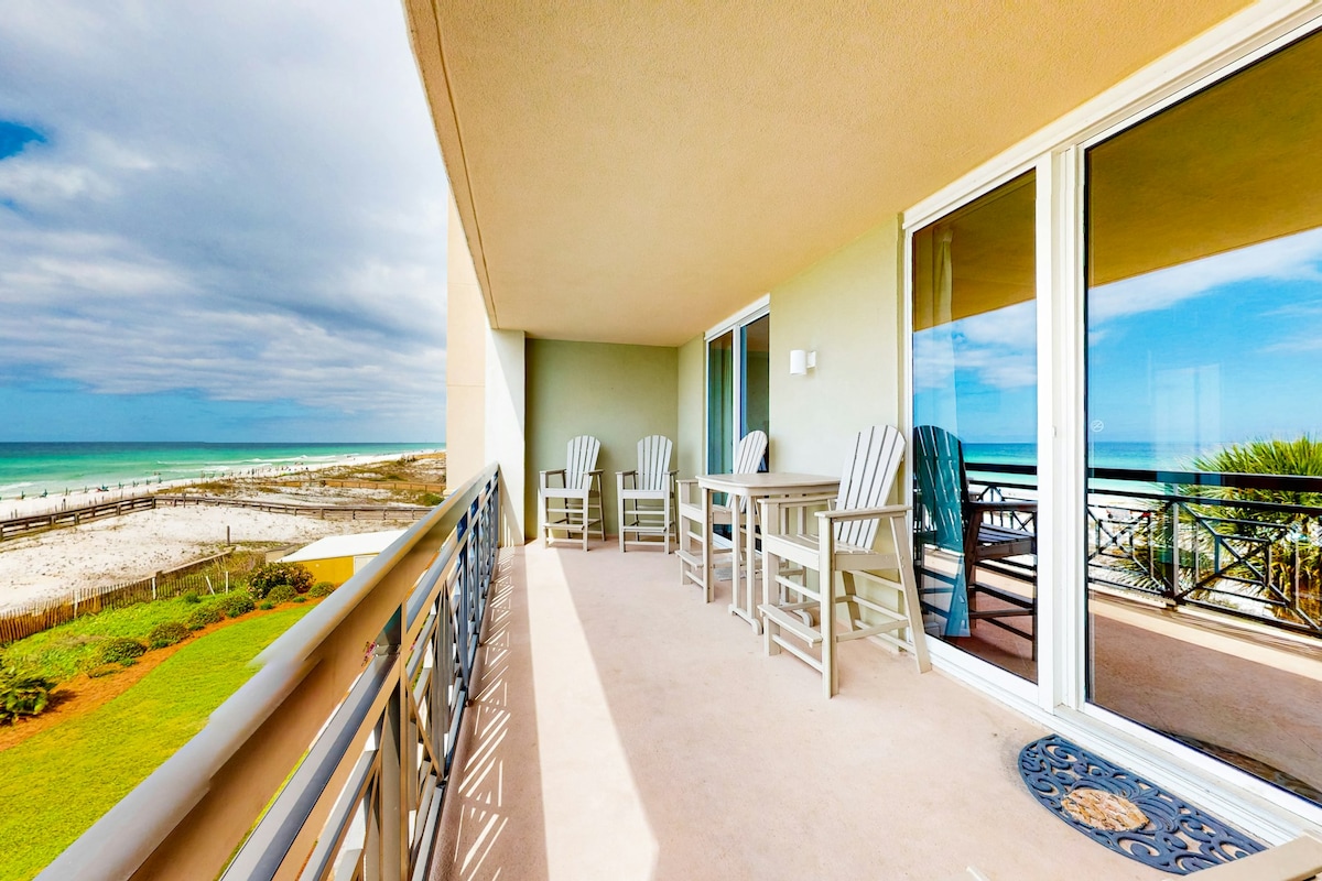 A spacious balcony extends from the rental, featuring several outdoor seating options. The view reveals the shimmering Gulf of Mexico, while soft, sandy beaches are visible in the distance under a partly cloudy sky.