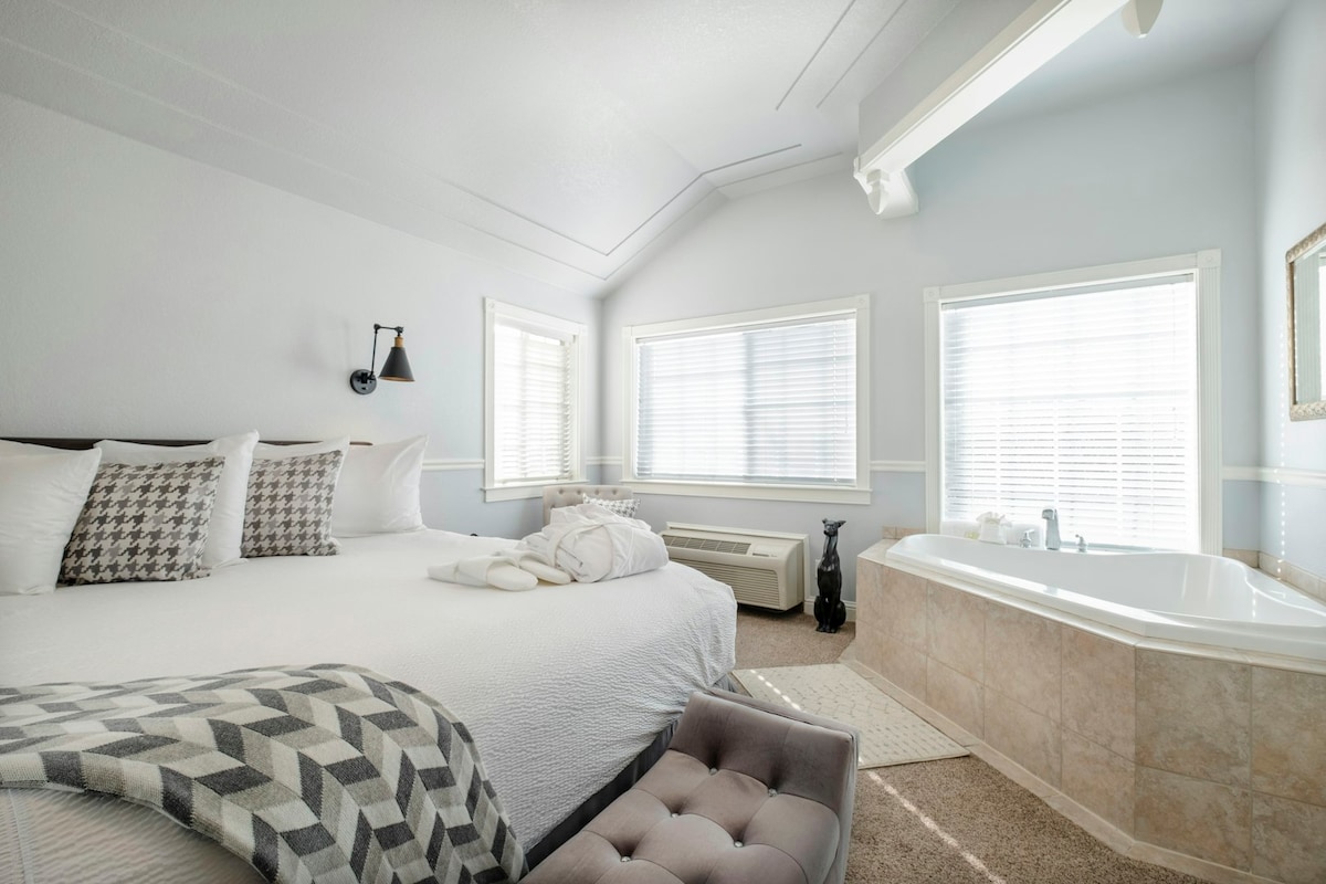 A spacious bedroom features a large bed adorned with decorative pillows and a cozy blanket. A relaxing seating area is positioned nearby, with natural light filtering through multiple windows. A whirlpool tub is located in one corner, complemented by soft, neutral furnishings.