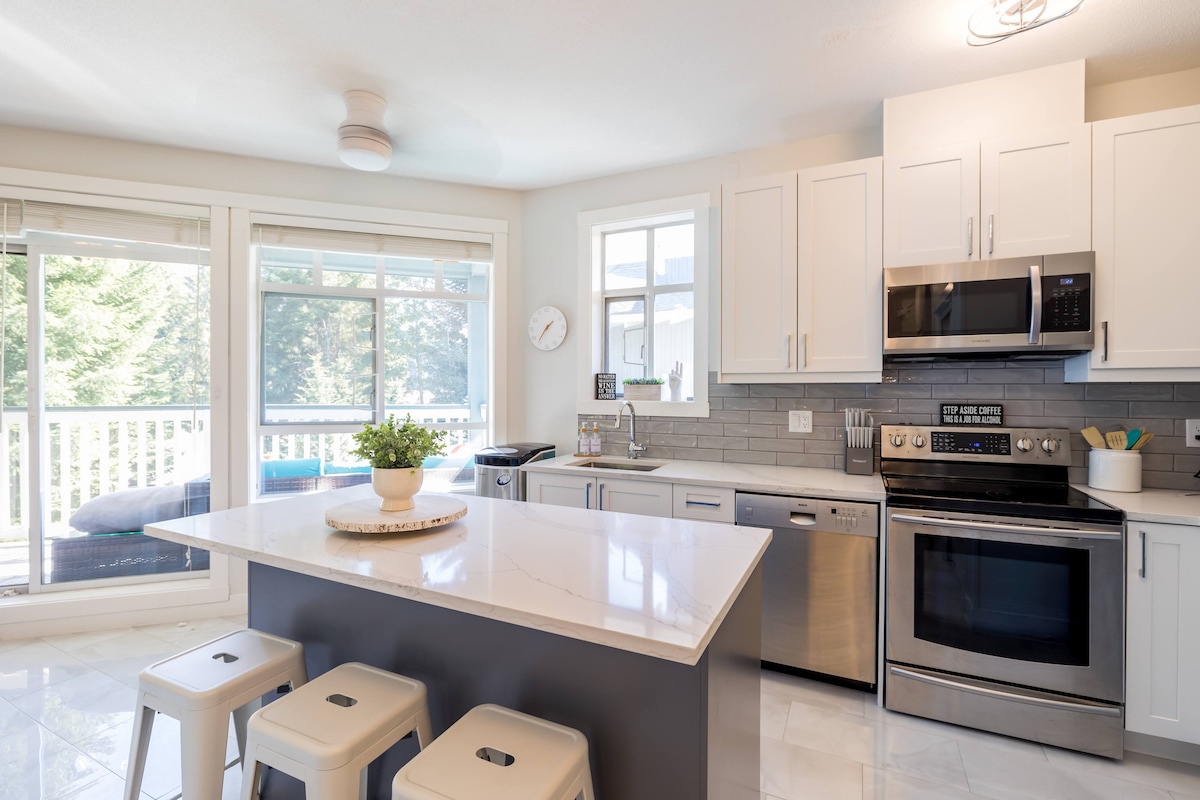A modern kitchen features stainless steel appliances and sleek cabinetry, complemented by a large island with seating. Natural light floods through multiple windows, with a view of the outdoor deck visible. A small potted plant adds a touch of greenery to the countertop.