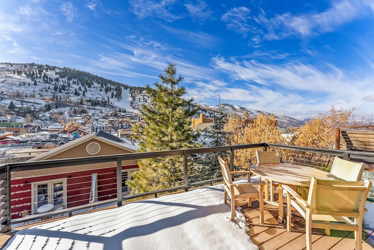 A spacious deck is featuring outdoor seating with a table and chairs, offering sweeping views of downtown Park City and the surrounding hills. A snowy landscape creates a serene backdrop beneath a bright blue sky scattered with wispy clouds.
