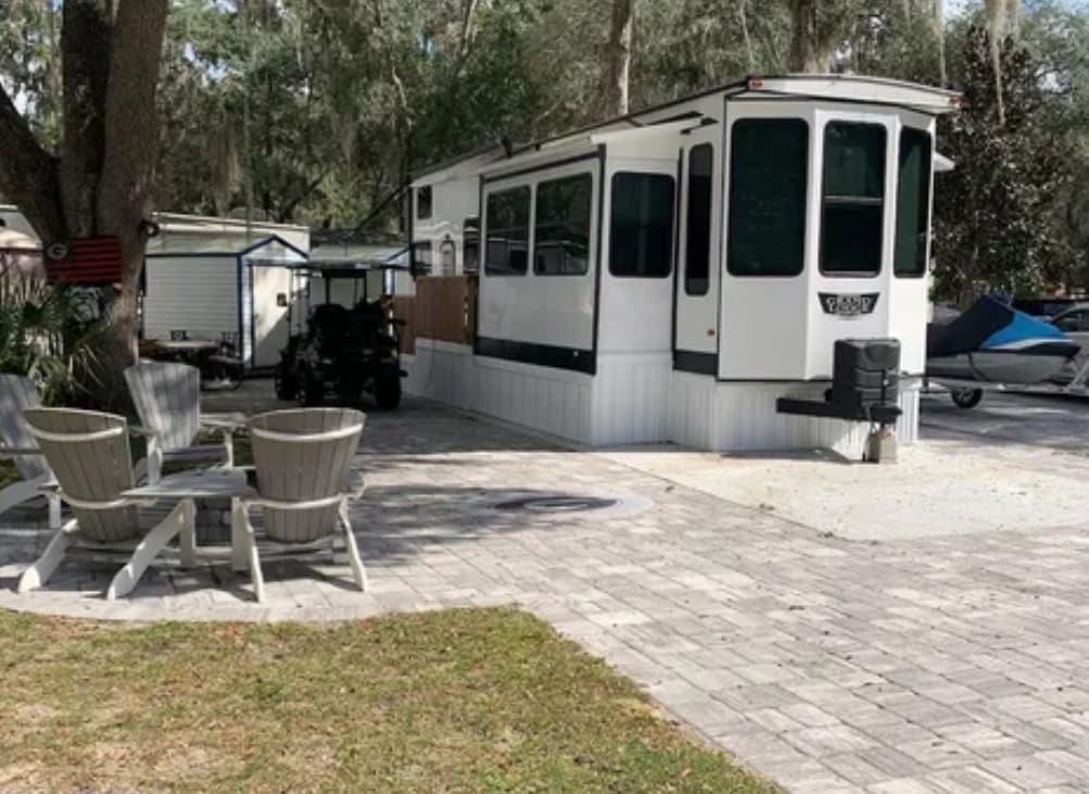 An RV is positioned on a paved area, complemented by a lawn of green grass. Two wooden chairs are arranged around a small outdoor space, creating a casual seating area. Nearby, a golf cart is visible, enhancing the convenience of mobility within the resort.