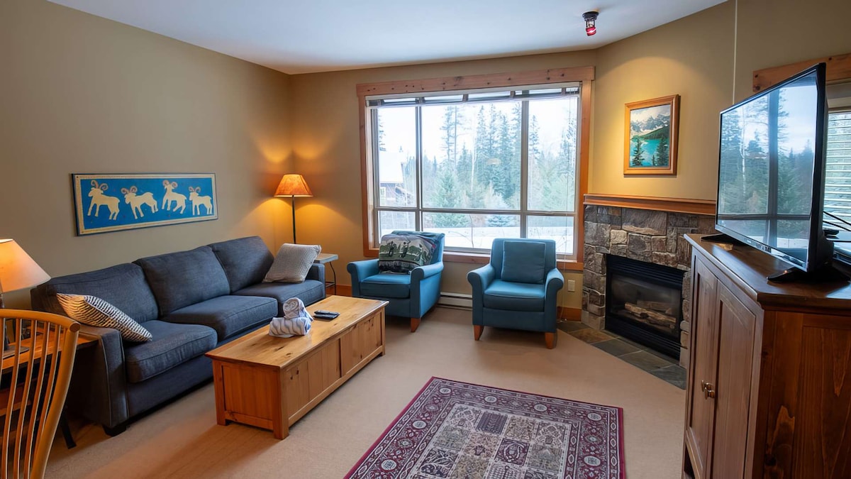 The living area features a comfortable gray sofa, two blue armchairs, and a wooden coffee table. Natural light fills the space from a large window showcasing views of the surrounding trees. A stone fireplace adds warmth, and a television is positioned for easy viewing.