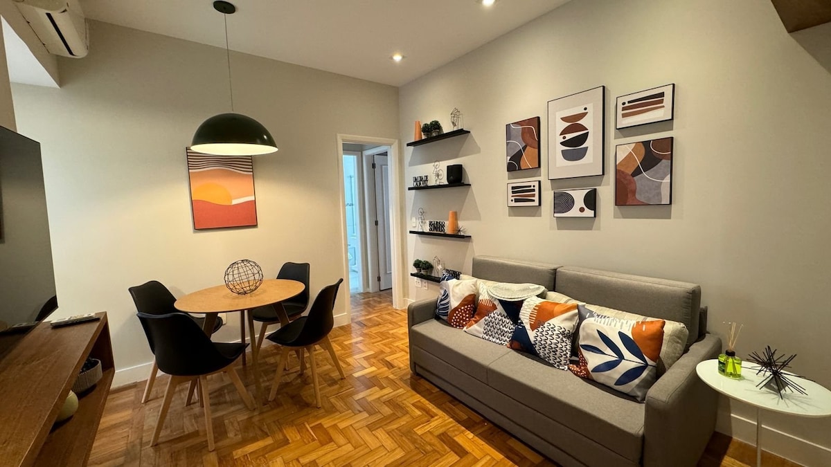 A comfortable living area is featured, showcasing a gray sofa adorned with decorative cushions. A round dining table with four black chairs is positioned nearby. Wall shelves display various decorative items, and framed artwork adds color to the light walls. Natural light illuminates the space.
