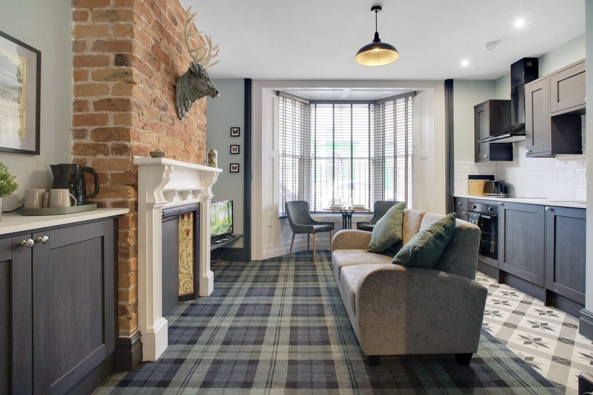 A cozy living area features a comfortable sofa with accent pillows, facing a wall-mounted TV. A decorative fireplace is flanked by contrasting dark cabinets. Large windows allow natural light to fill the space, complemented by stylish plaid carpeting and a charming dining table for two.