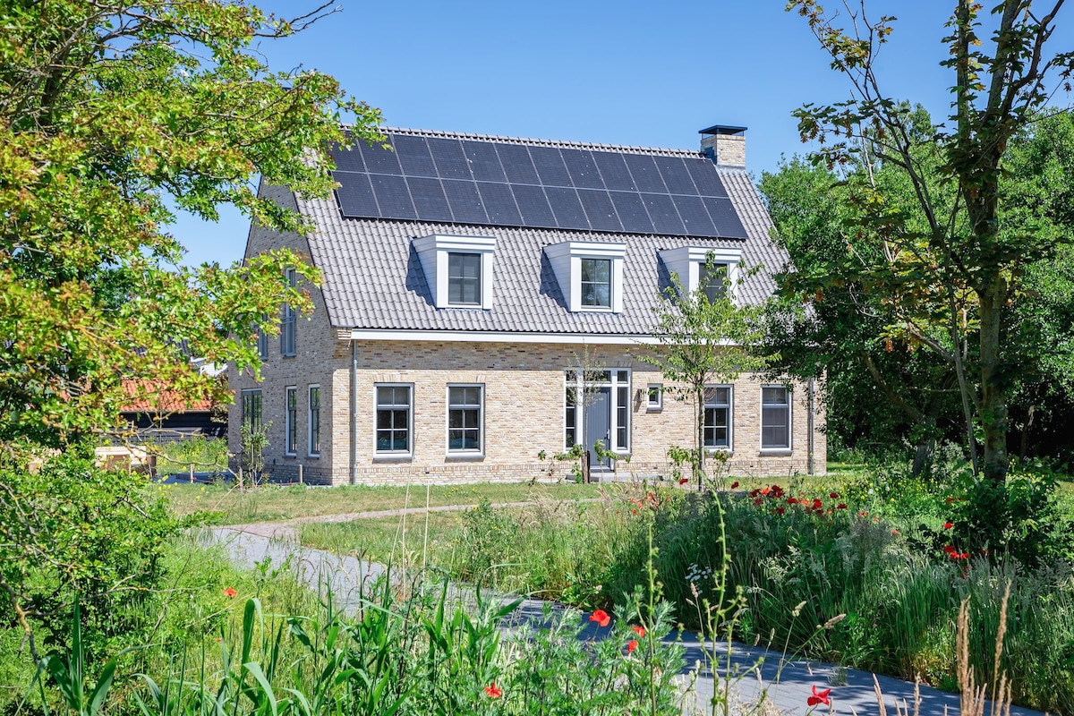 A spacious family villa is displayed, featuring a light-colored stone exterior and a slate roof equipped with solar panels. Surrounding the house, a vibrant garden is filled with wildflowers, greenery, and pathways leading towards the entrance.