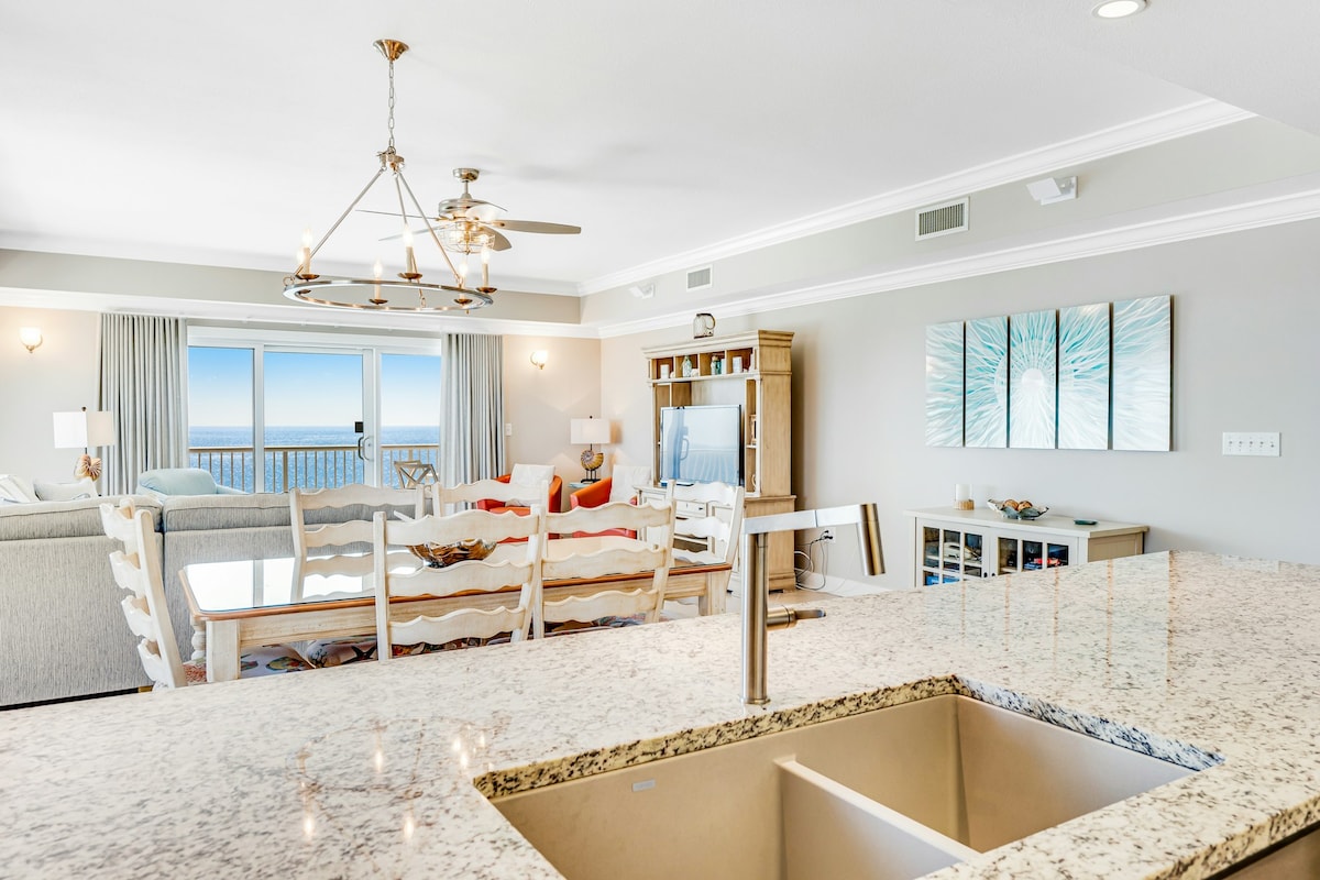 An open-concept living area is visible, featuring a spacious kitchen island with a sleek sink. The dining area includes a long, wooden table with several white chairs. Bright windows offer a view of the gulf, while a cozy seating area is located beyond, adorned with light-colored furnishings.