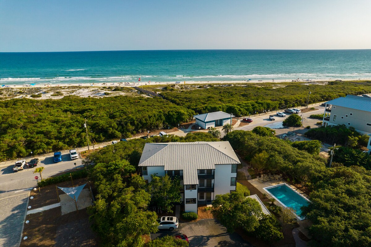 Aerial view captures the coastal community with lush greenery surrounding a two-story condominium. The sparkling ocean waves and sandy beach are visible in the distance, framed by the natural landscape. A community pool sits nearby, enhancing the tranquil atmosphere.