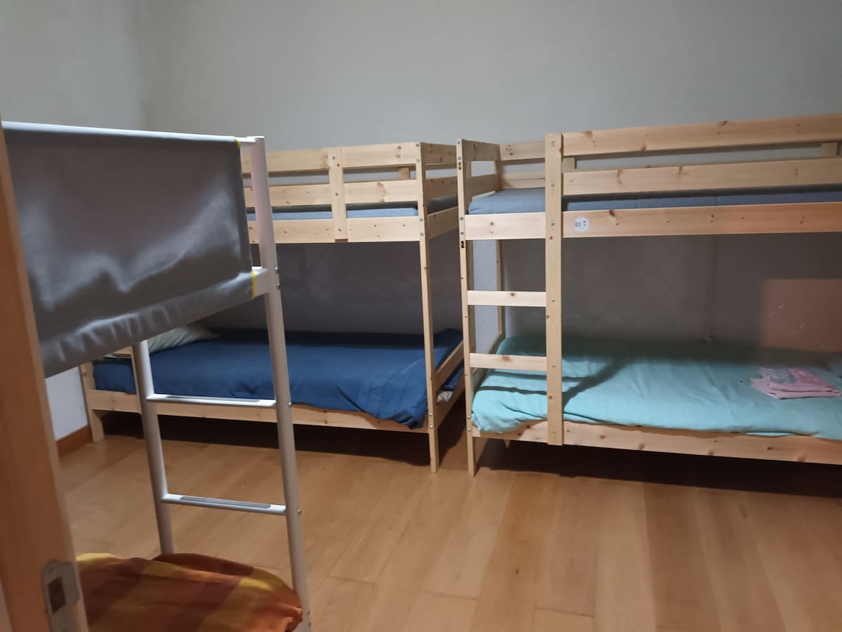 A shared dormitory room features three wooden bunk beds, each with single mattresses in solid colors. Wooden floors create a warm atmosphere, while the simple layout allows for easy movement. A small table is visible, and a door leads to the room's entrance.