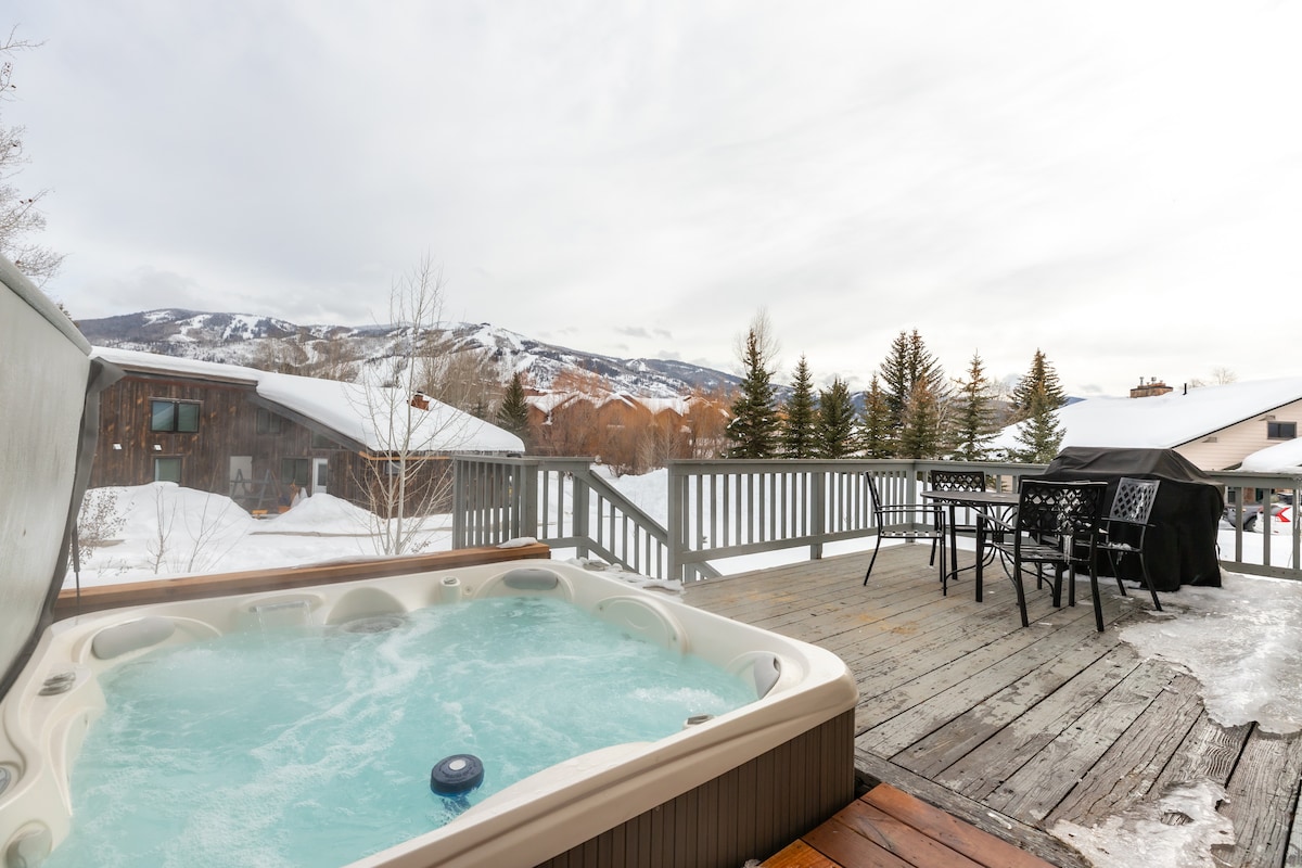 A hot tub is positioned on a wooden deck, offering panoramic views of the snow-covered mountains. A dining set with chairs and a grill is arranged nearby, providing outdoor space for relaxation and gatherings amidst the winter scenery.