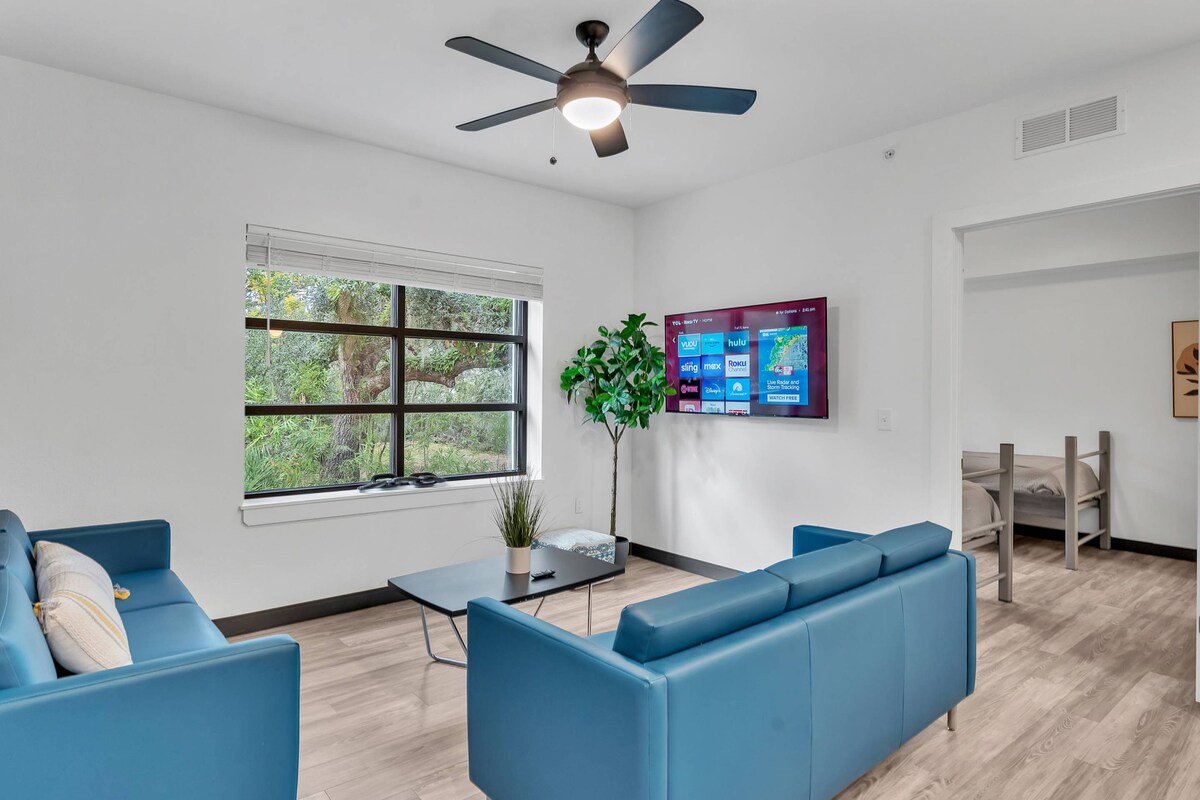 A bright living area is furnished with two blue sofas facing a coffee table. A ceiling fan hangs above, and a large window allows natural light to fill the space while providing a view of greenery outside. A wall-mounted Roku Smart TV is positioned for easy viewing.