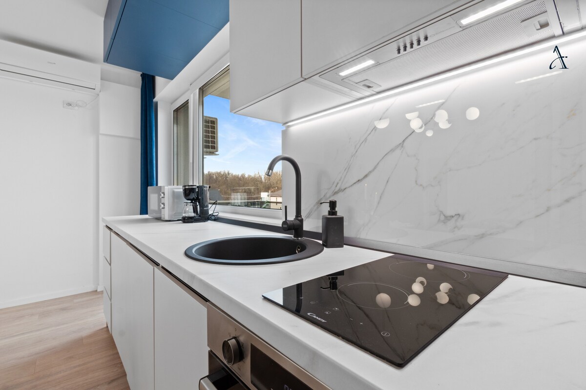 A modern kitchen is displayed with a sleek silhouette, featuring a black sink and induction cooktop. White cabinetry contrasts with a marble backsplash, illuminated by natural light from the window. Essential appliances, including a microwave, are neatly arranged, enhancing the space's functionality.