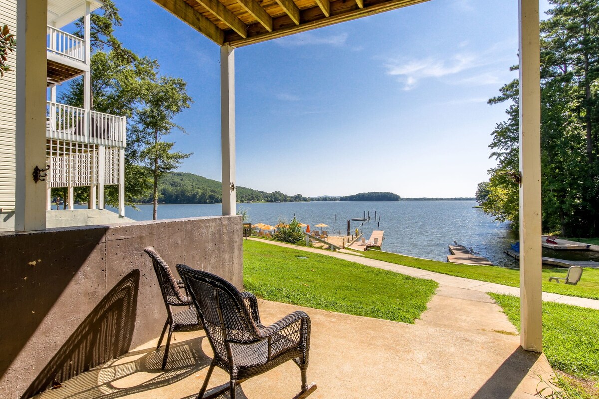 A covered porch area features two black wicker chairs facing an expansive lake view. The lush green lawn leads directly to the water, with a private dock visible in the distance. Mountains frame the horizon under a clear blue sky.