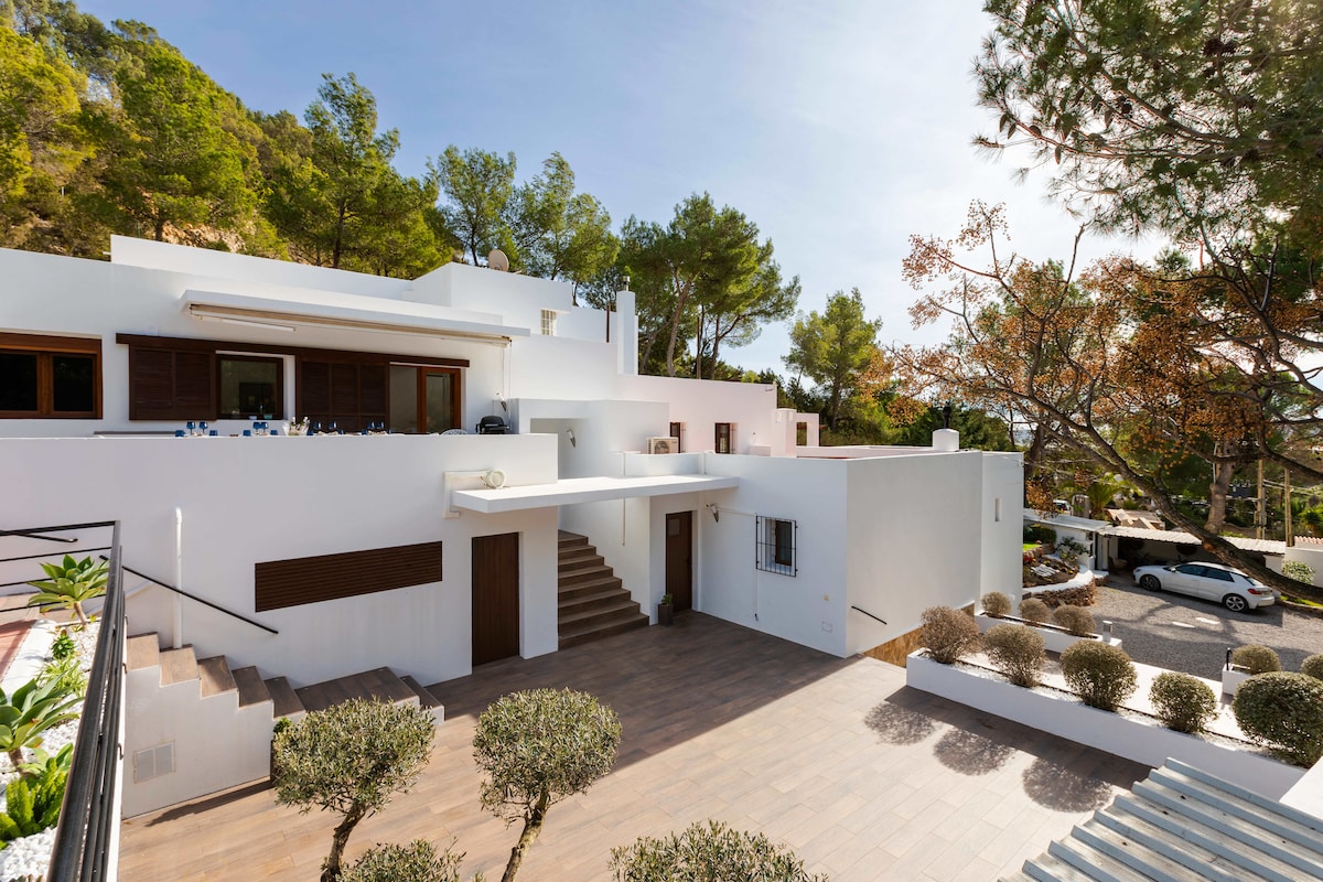 A modern villa is presented, showcasing a clean white exterior surrounded by lush greenery. Stone pathways lead to the entrance, and steps ascend to the upper level. Outdoor spaces feature manicured shrubs, providing a serene atmosphere in a secluded setting.