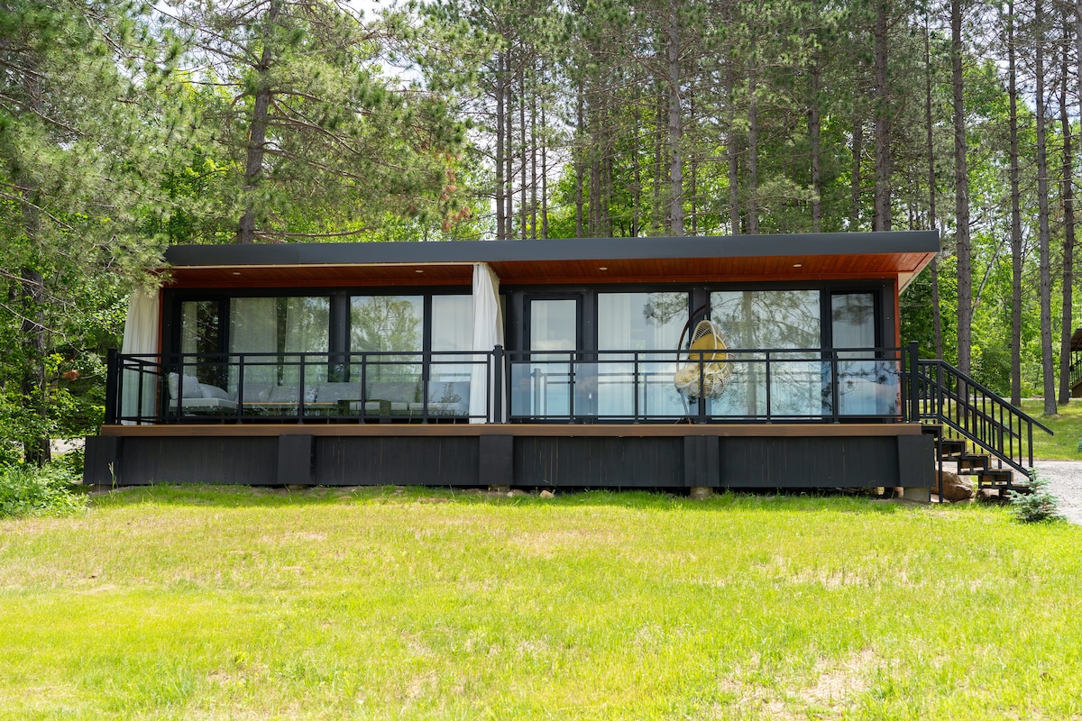 The cottage exterior displays a modern design characterized by large glass windows and wooden paneling. A spacious deck is found at the front, featuring a railing that provides a clear view of the surrounding greenery, and highlighting the connection to the natural landscape.