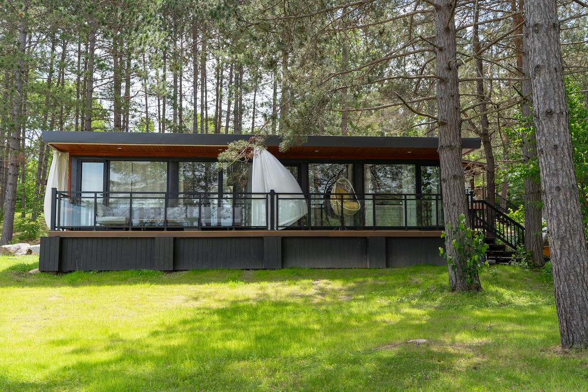 A modern cottage is framed by lush greenery and towering trees. Its all-glass front is complemented by a spacious wooden deck, which features a hanging chair. Natural light floods the interior through the expansive windows, creating a seamless connection with the outdoors.