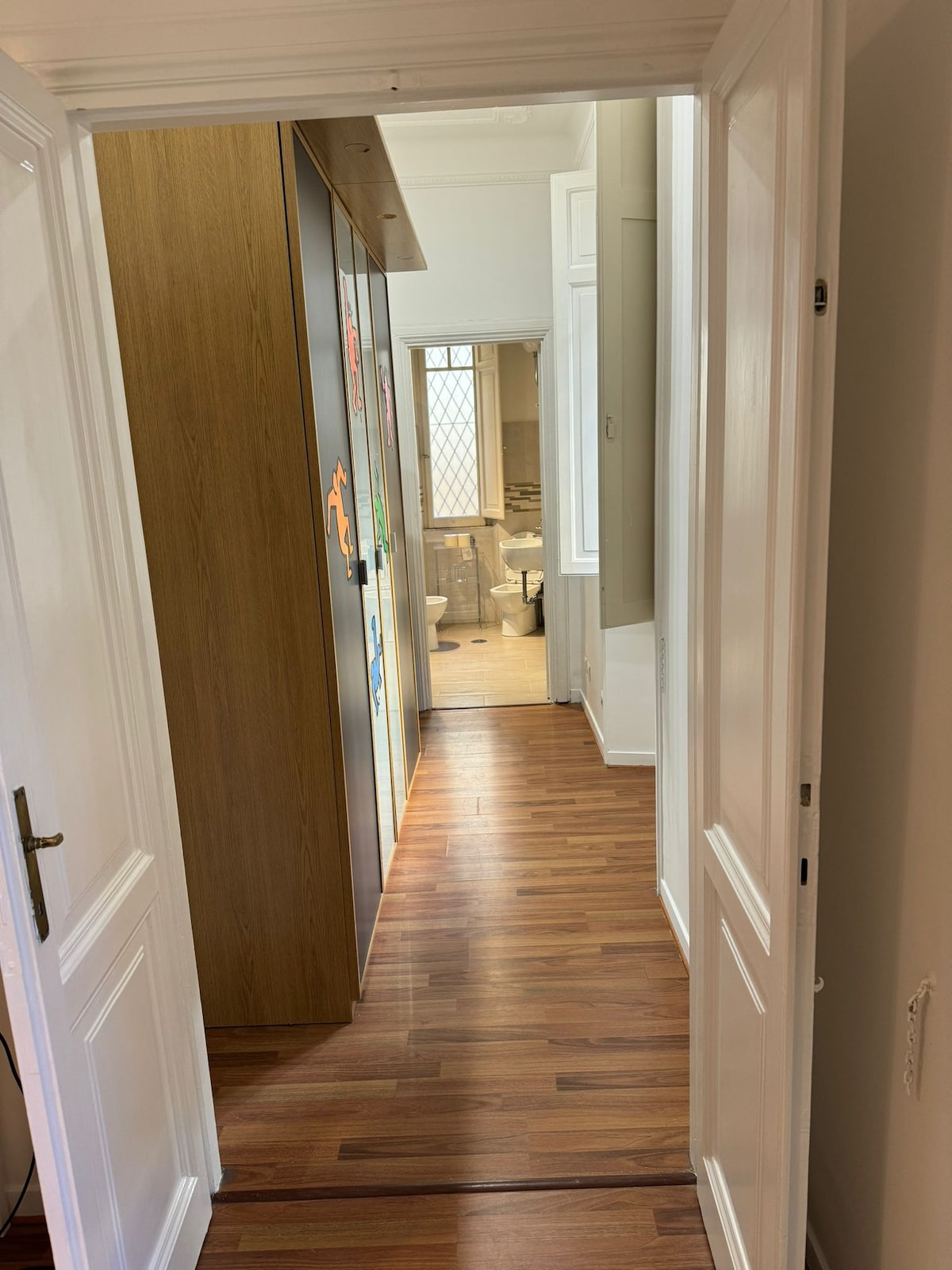 A hallway featuring wooden flooring and light-colored walls is shown. Storage cabinets line one side, while a doorway at the far end leads to a bathroom area. Natural light filters in through a window, creating a warm and bright ambiance.