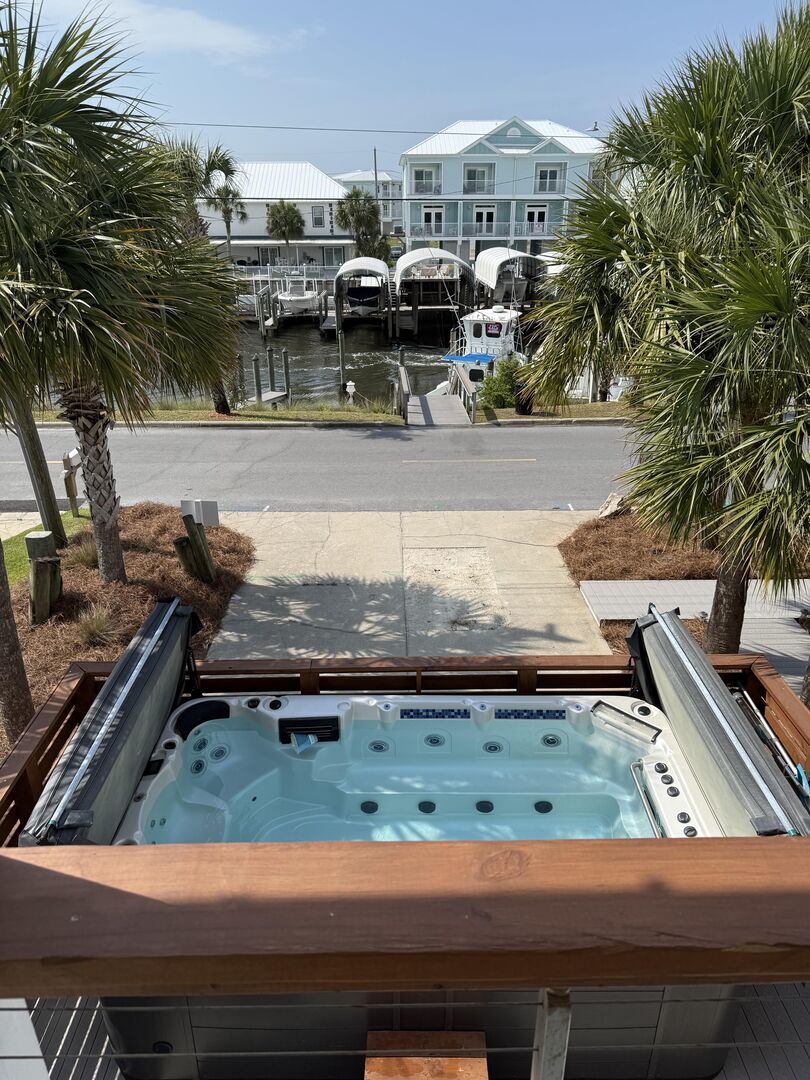An oversized, heated swim spa is prominently featured on the deck, surrounded by a wooden railing. Lush palm trees frame the view, overlooking a canal with boats docked nearby. A clear blue sky provides a bright backdrop, enhancing the outdoor relaxation space.