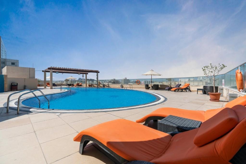 An outdoor swimming pool is surrounded by lounge chairs with orange cushions. A shaded pergola stands nearby, offering a relaxing space. Clear blue skies and a few sun umbrellas enhance the comfort of the area, creating a serene setting for leisure and relaxation.