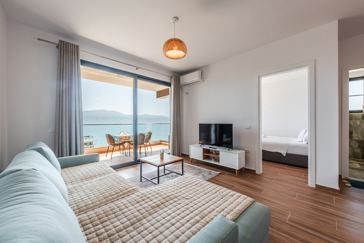 A spacious living area is furnished with a light-colored couch and a coffee table, facing large glass doors that provide an unobstructed view of the sea. A flat-screen television sits on a minimalist console. A door leads to a softly lit bedroom in the background.