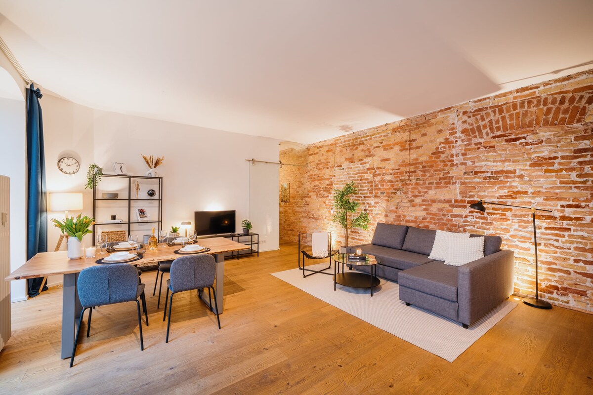An open living space features a stylish gray sofa and a wooden dining table set for four. A warm toned brick wall adds texture, while a smart TV is positioned on a low stand. Potted plants and decorative items enhance the room's inviting feel.