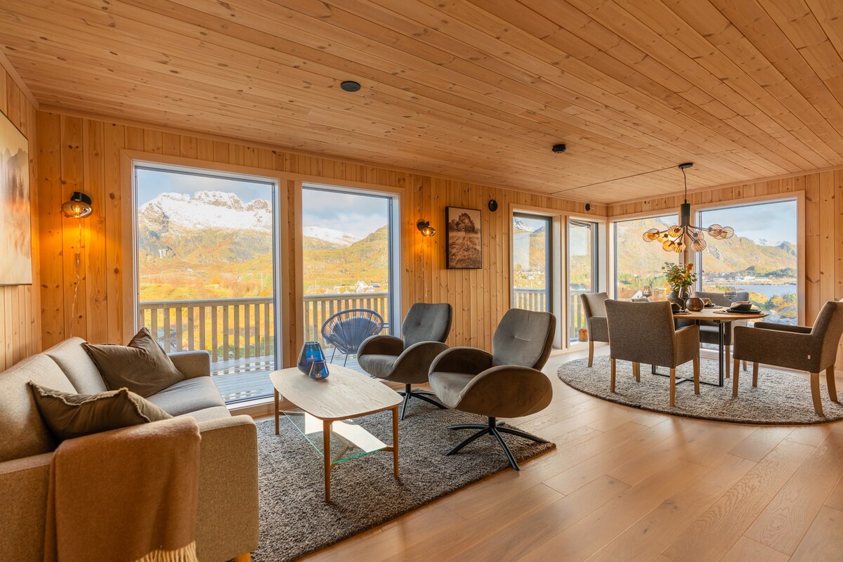 A spacious living area features natural wood paneling and multiple large windows offering scenic views. Comfortable seating arrangements include a sofa and stylish chairs, complemented by a round dining table. Soft lighting enhances the inviting atmosphere, while a textured rug anchors the seating area.