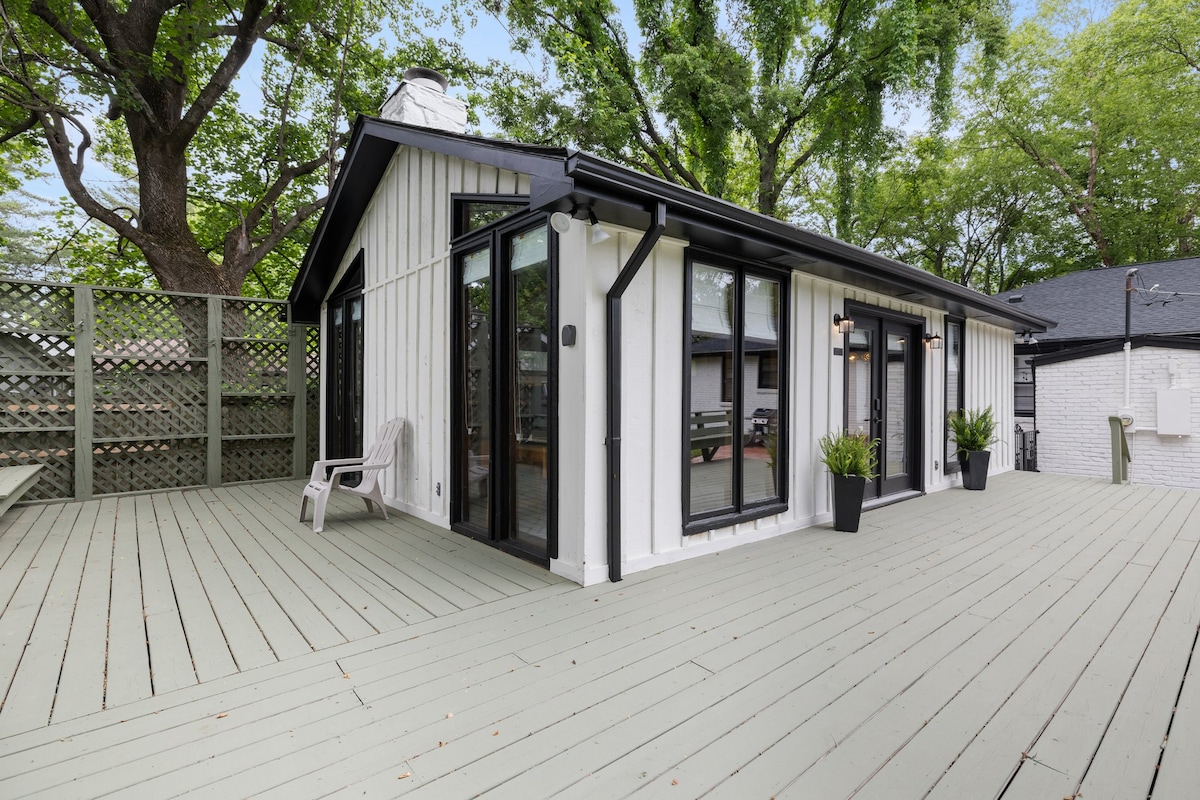 A spacious outdoor deck is surrounded by lush greenery and features a light-colored wooden floor. Large glass doors provide access to the interior, while potted plants and seating options add to the inviting atmosphere of the area.