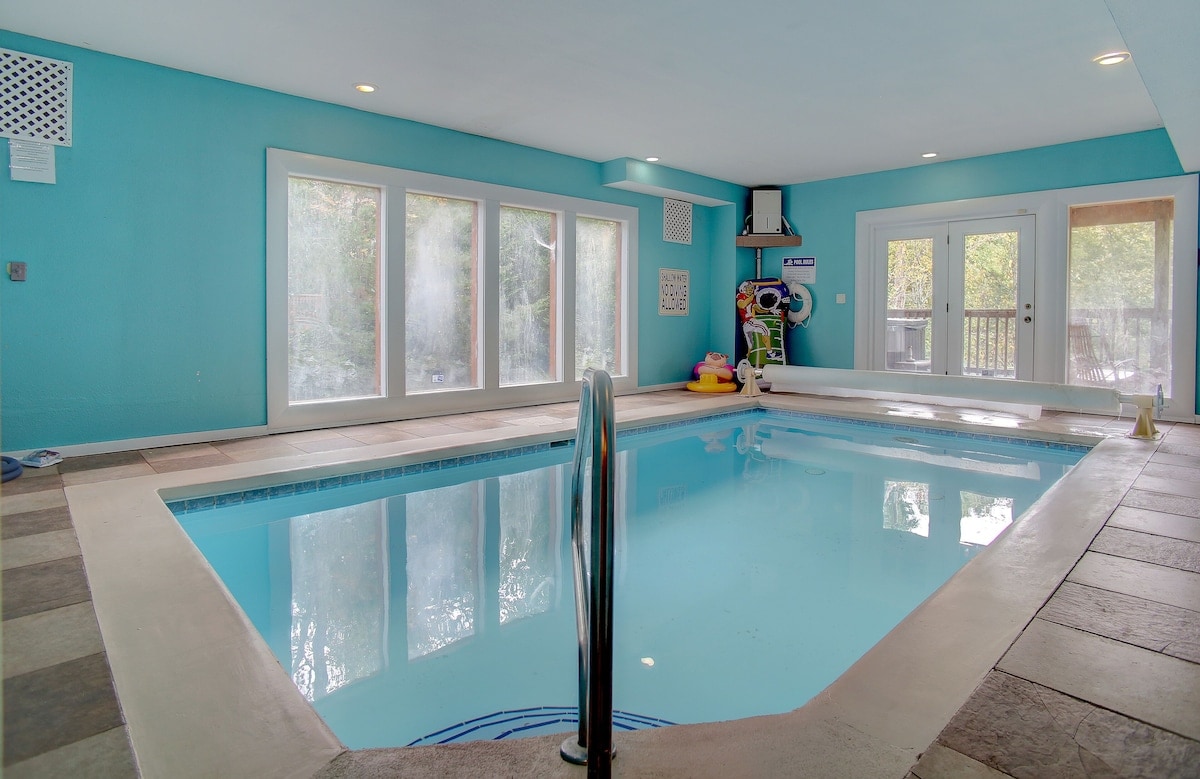An indoor heated pool is surrounded by bright blue walls, creating a cheerful atmosphere. Large windows allow natural light to fill the space, with a view of the outdoors. A railing enhances safety, and colorful pool toys can be seen nearby.