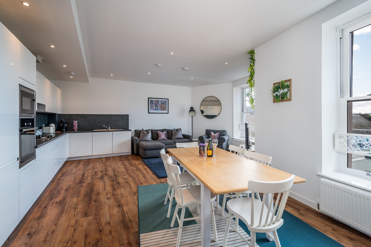 A modern open-plan living area is presented, featuring a stylish kitchen with sleek white cabinetry. A light wood dining table with white chairs is positioned in the center, while a comfortable grey sofa and a rug define the relaxation space. Large windows allow natural light to fill the room.