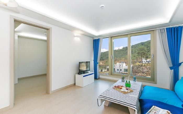 A bright living area features large windows offering views of the surrounding landscape. A cozy sofa is positioned alongside a low table set with refreshments. Nearby, a television stands on a console, and soft lighting enhances the open, airy feel of the space.