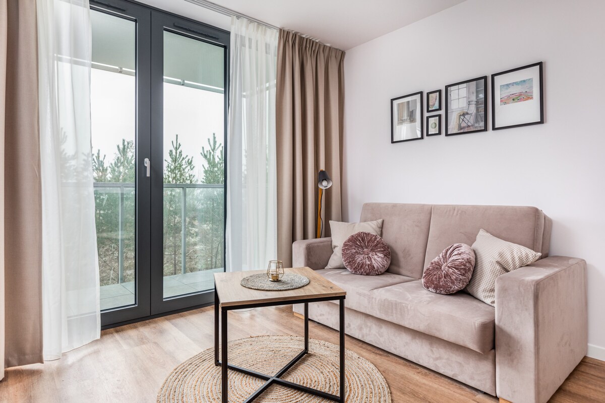 The living area features a comfortable fold-out sofa adorned with cushions. A coffee table sits in front, complemented by a round rug on the wooden floor. Large glass doors open to a balcony, framed by soft, light-colored curtains.