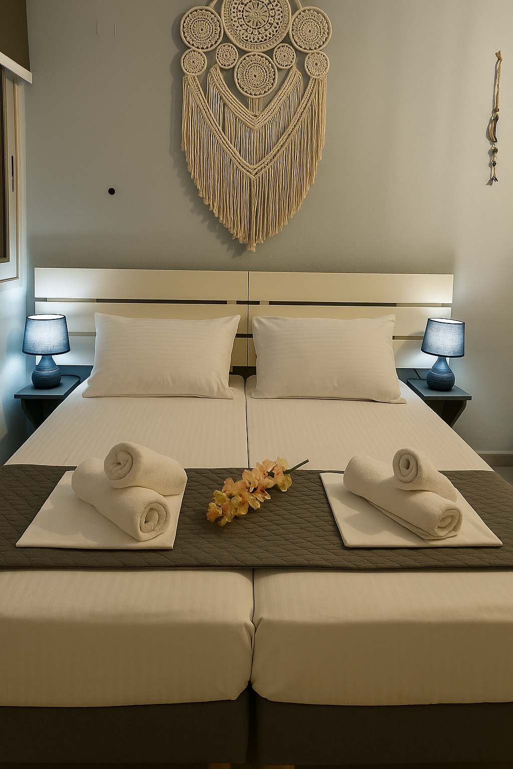 A calm sleeping area features two single beds that can be combined into a double, dressed in crisp white linens. Soft lighting is provided by two bedside lamps, enhancing the tranquil ambiance. A decorative wall hanging adds a touch of texture above the beds, complemented by neatly arranged towels.