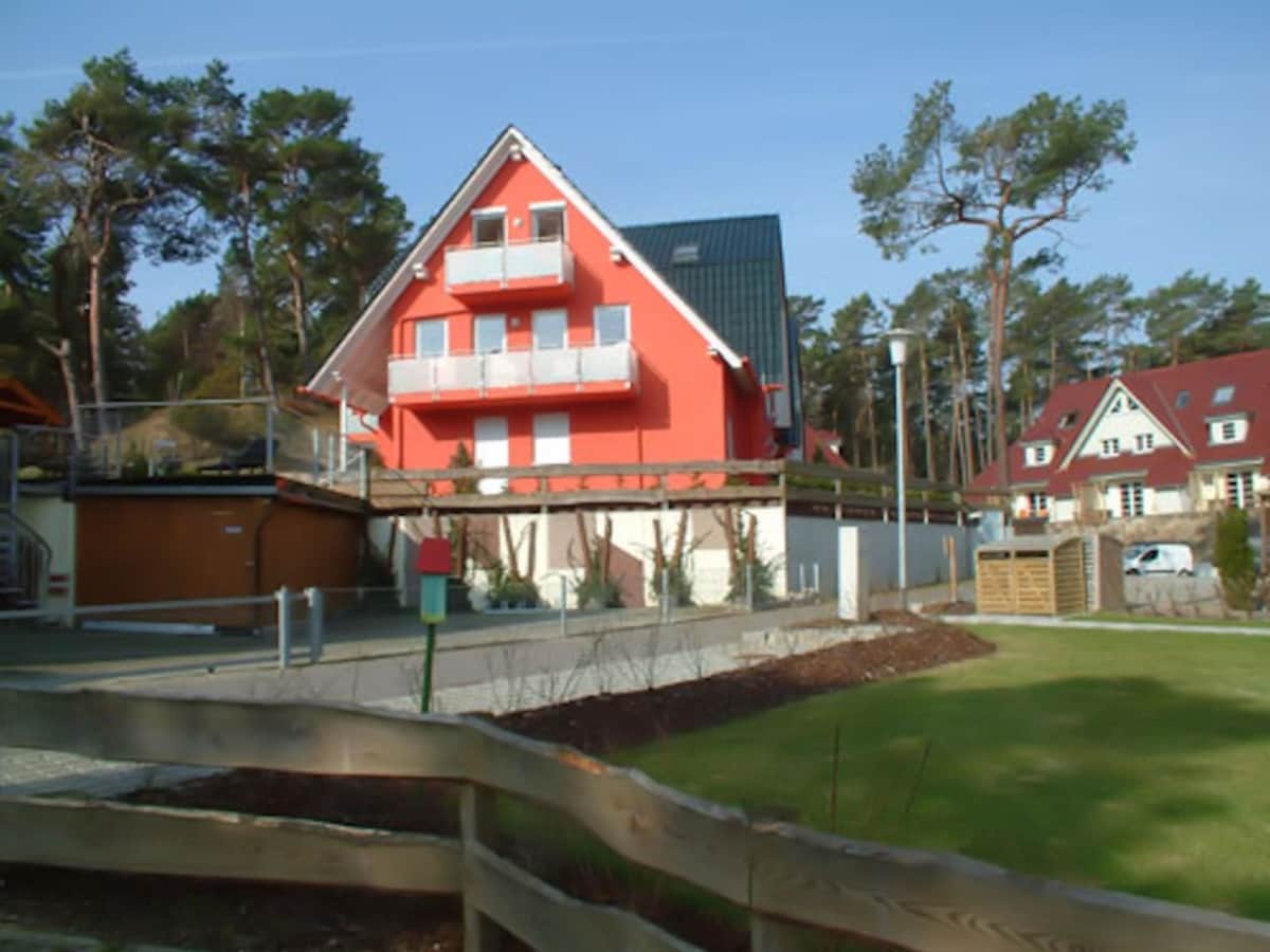 A two-story red and gray building features multiple balconies surrounded by trees. The exterior is complemented by landscaped greenery and wooden fencing, with pathways leading up to the entrance, providing a welcoming appearance.