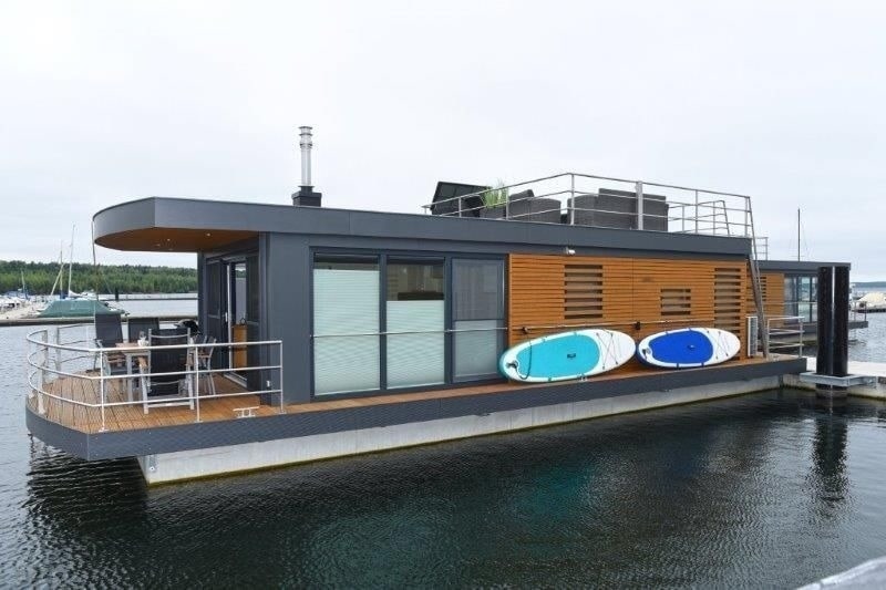 The houseboat features a modern design, with a spacious deck adorned with two paddleboards. Large glass windows provide ample light to the interior, while the stylish exterior blends sympathetically with the surrounding water.