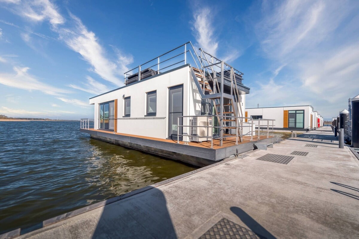 A modern floating house is anchored on the water, featuring a significant deck leading to an upper level. The structure displays large windows that offer expansive views, and a stairway connects the two levels. Calm waters surround the house, enhancing its tranquil setting.