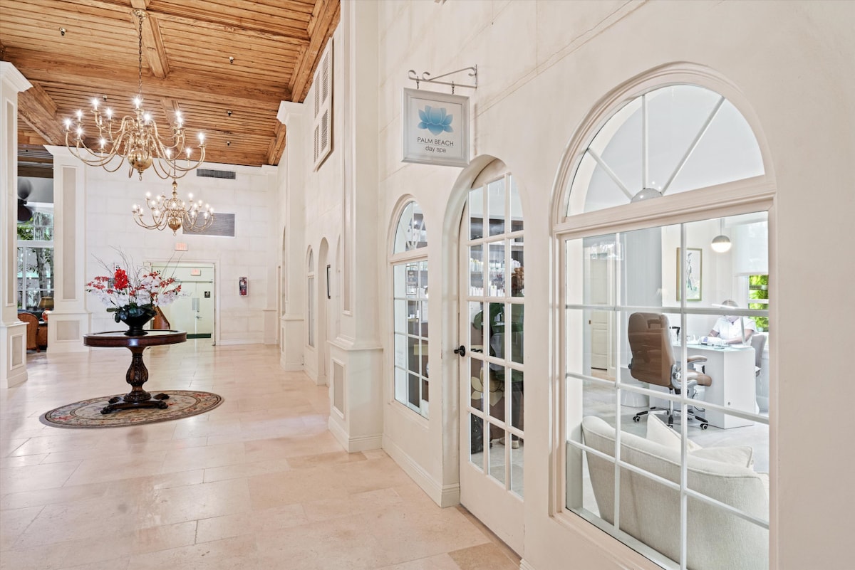 The spacious lobby features high ceilings with wooden beams and elegant chandeliers. Large arched windows allow natural light to flood in, framing a modern seating area with a chic armchair and desk. Decorative floral arrangements add a touch of warmth to the entrance.