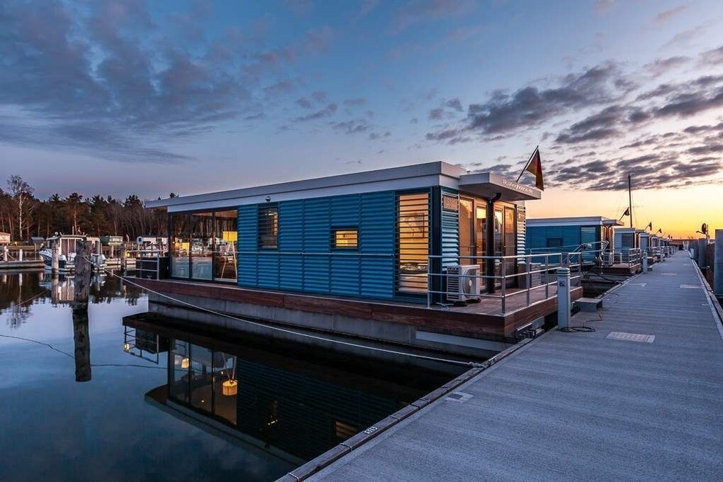 A floating house is anchored along a calm marina at dusk, featuring blue exterior siding and large windows. Soft reflections can be seen on the water's surface, while the surrounding area is illuminated by warm light from within the house.
