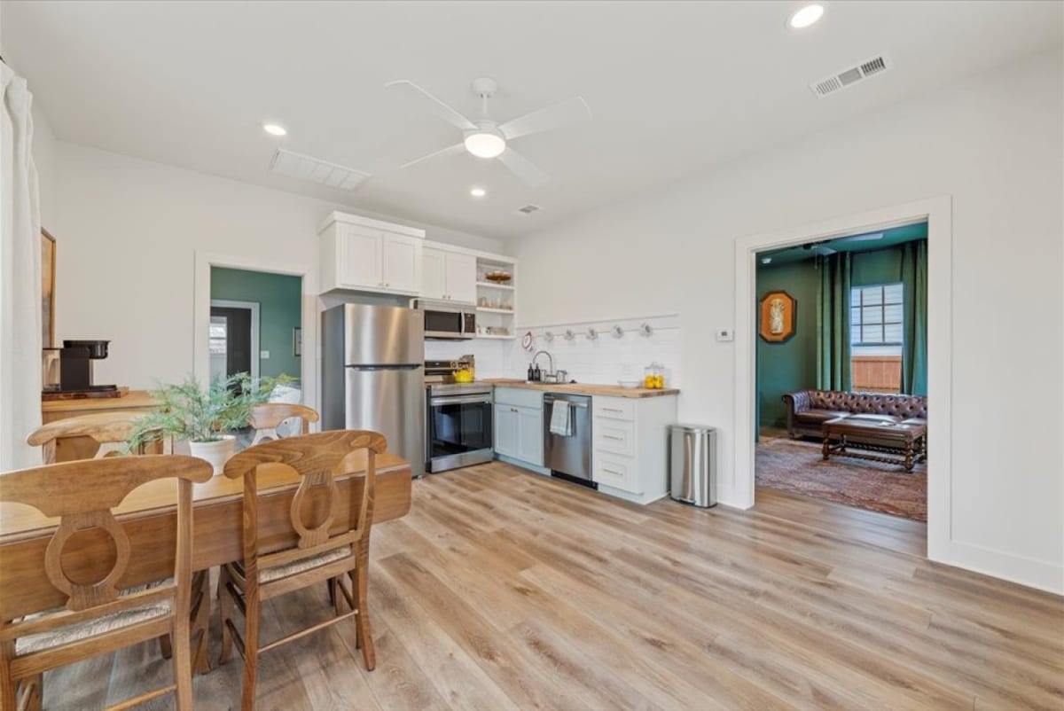 A modern kitchen and dining area feature a wooden dining table with four chairs and stainless steel appliances. A ceiling fan is visible above, and a doorway leads to a sitting room with rich green walls and a window framed by light curtains.
