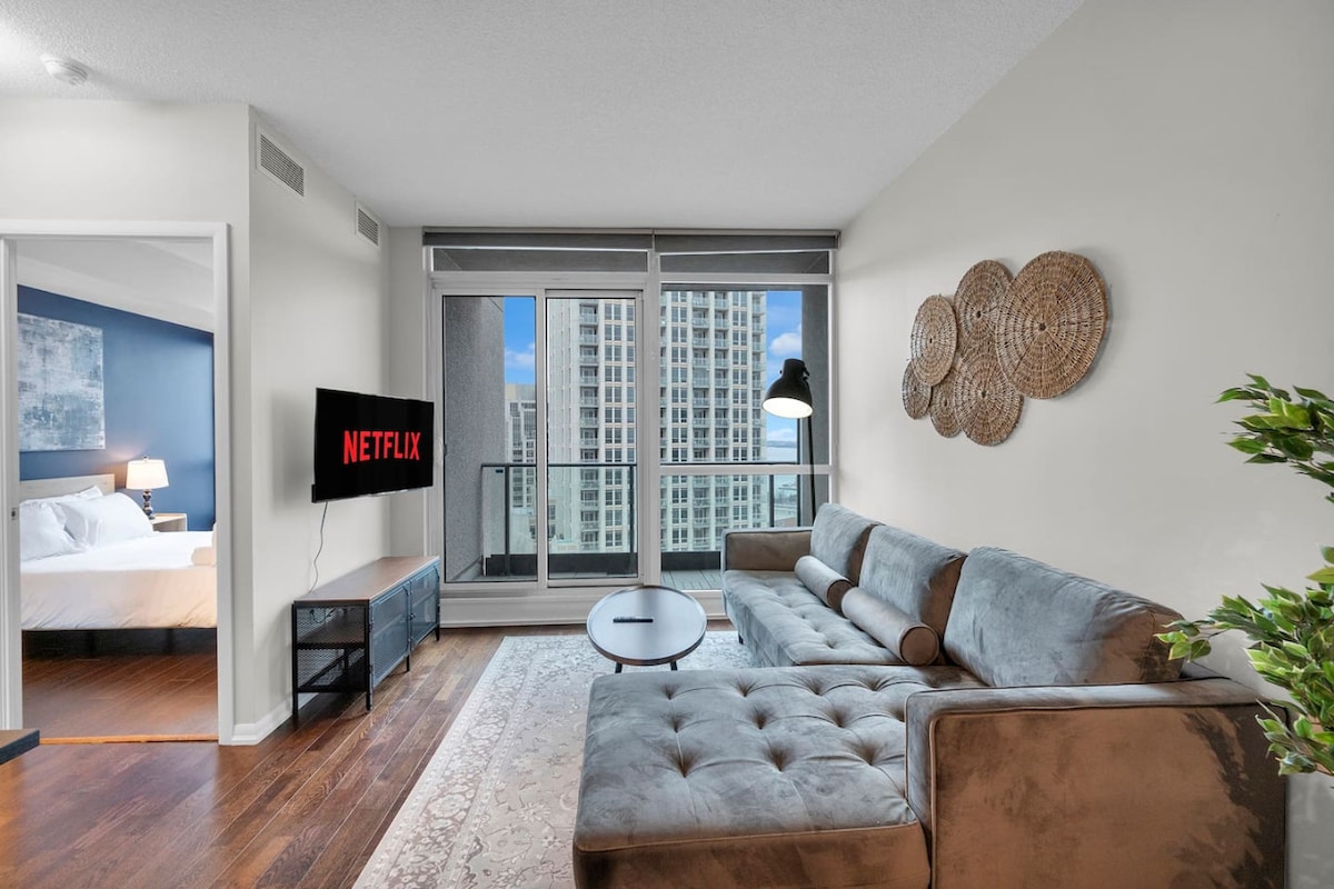 A modern living area is showcased, featuring a plush gray sectional sofa and a round coffee table. A wall-mounted television displays the Netflix logo. Large windows provide natural light and a glimpse of the city skyline. The space is complemented by decorative wall hangings.