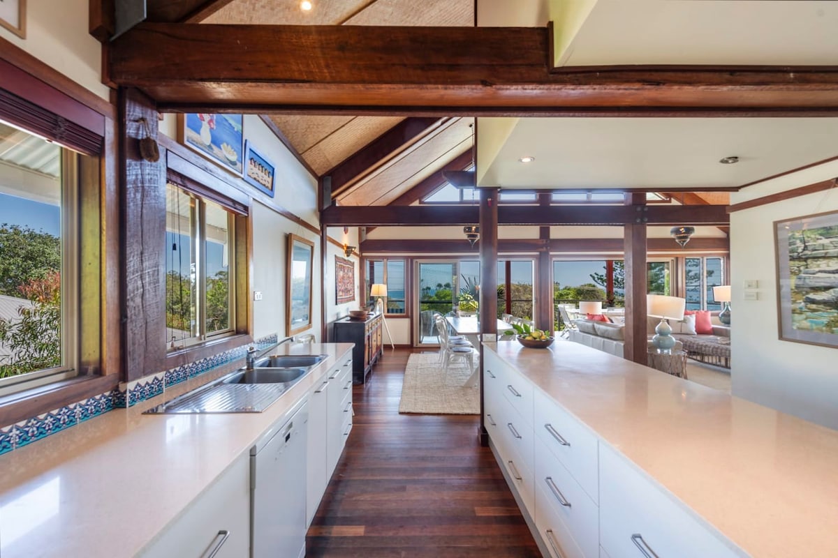 The kitchen features expansive countertops and modern appliances, with large windows that provide natural light and views of the surrounding landscape. Timber beams and architectural details contribute to a spacious, open-plan design that connects seamlessly to the living area.