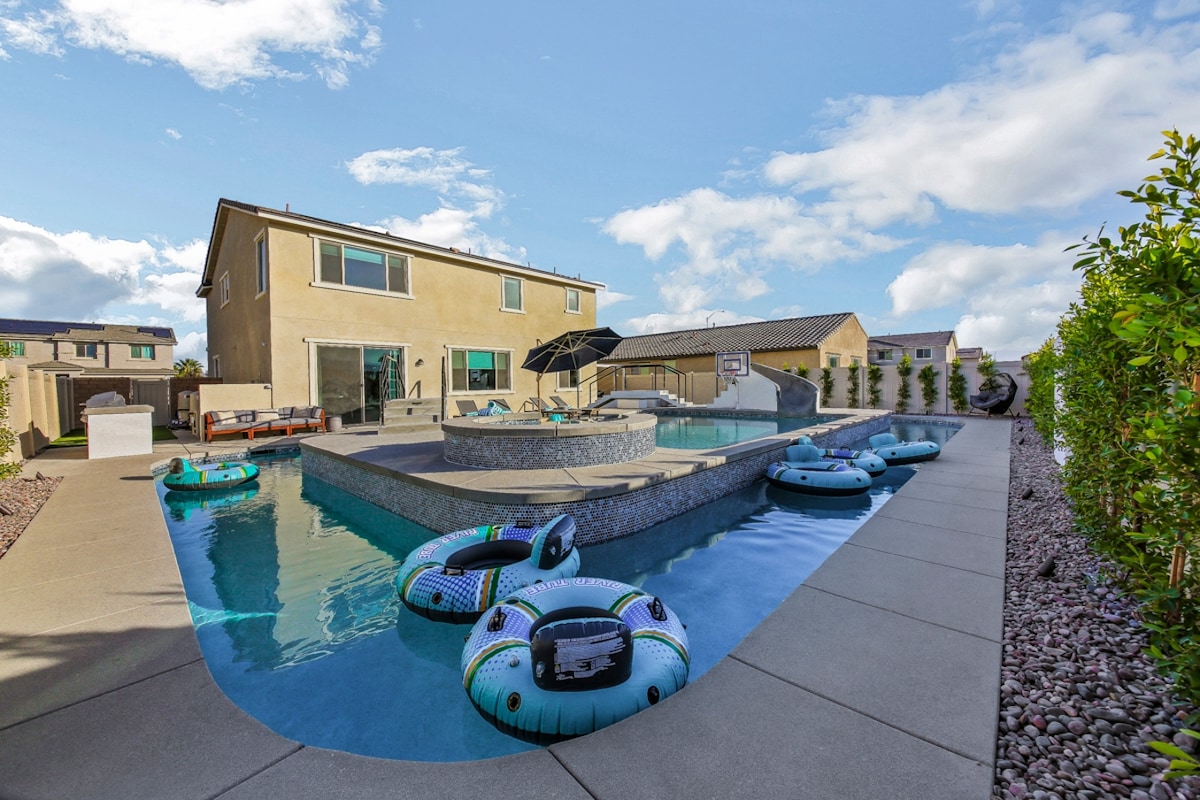 A sunny outdoor space features a winding lazy river flanked by seating areas and a grass-lined fence. Colorful inflatables float on the water's surface, while the home stands in the background with large windows showcasing the interior. A lounge area with umbrellas is visible nearby.